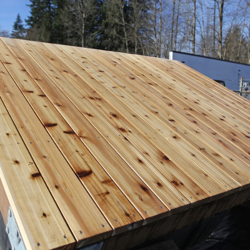A newly constructed, sloped wooden roof, shown from a low angle. The roof's natural woodgrain texture contrasts with a clear blue sky and bare trees.