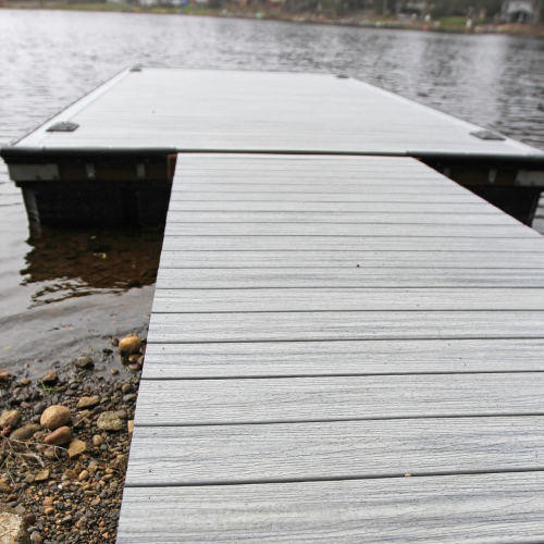A wooden dock extends over a calm lake, viewed from a rocky shoreline. The overcast sky creates a serene, peaceful atmosphere.