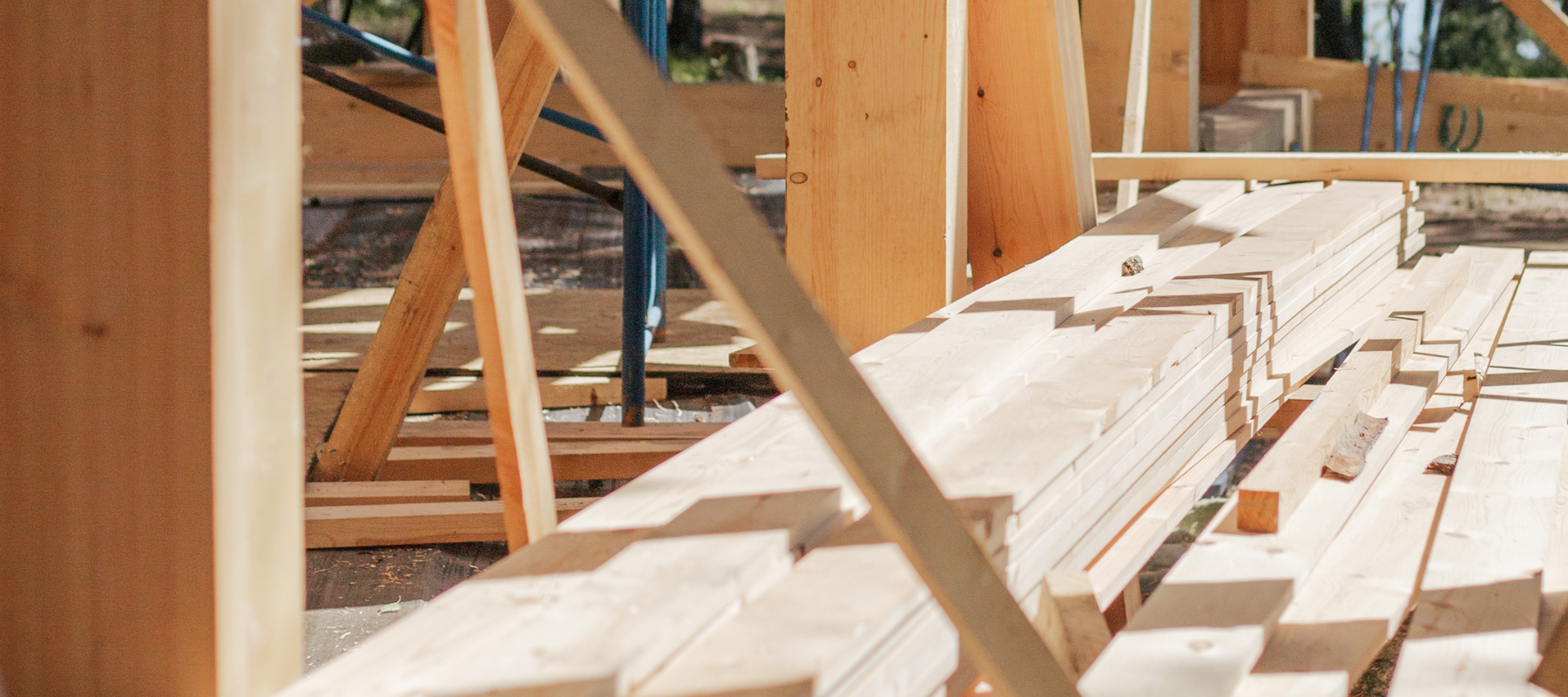 Stacks of light-colored wooden planks are arranged on a construction site. Wooden beams and scaffolding frame the scene, suggesting an active building project.